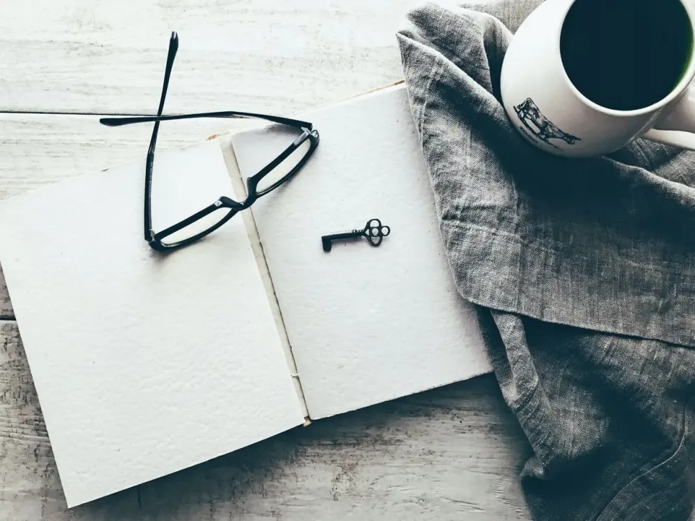 Looking down at an empty page in a notebook with key and glasses on top which are layin gon a wooden table
with a blanket next to it with a cup of coffee on top.